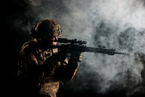 An army soldier in a military camouflage uniform, a helmet and a mask, holds a rifle and aims with a red dot sight, standing in an attack position, in the smoke on a black background.