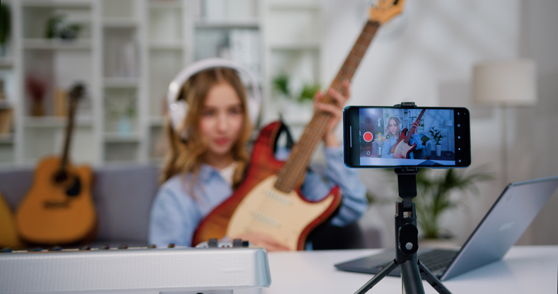 Blogger teen girl looking lesson recording, playing guitar. Cheerful female playing guitar using mobile phone. Online learning teaching education concept.