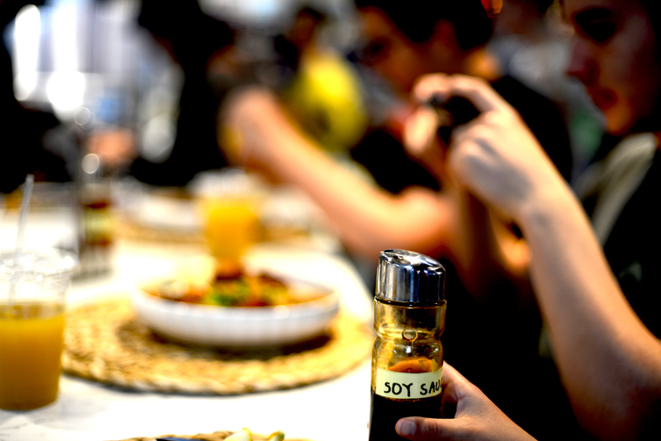 Close-up of Soy Sauce Container with Teenager Taking Photo of Meal in Blurred Background