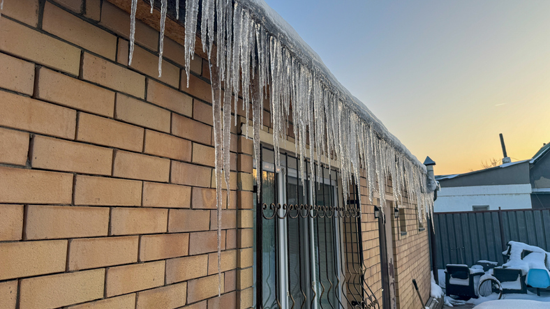 Long glistening icicles hanging from a building roof at sunset, reflecting warm golden light on a cold winter day.