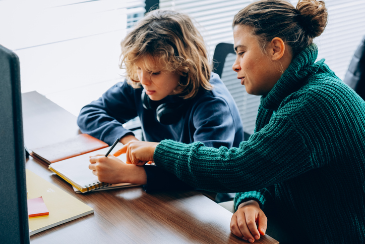 Young boy with curly blond hair writes in a notebook while a young woman in a green sweater watches attentively. Educational environment, tutoring, education support, learning process concept.