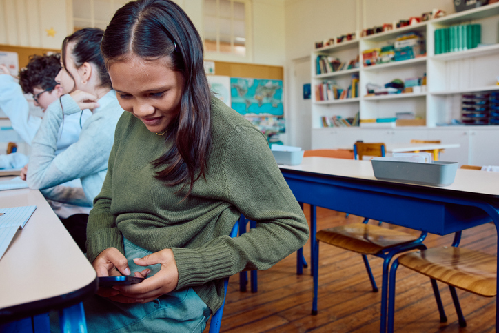Schoolgirl looking down and using her mobile phone under desk in classroom