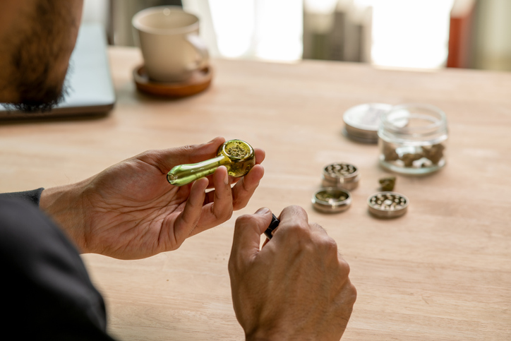Asian man smoking a Marijuana from smoking pipe, with cannabis equipment and accessories on the table.