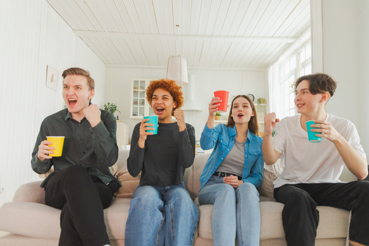 Group of friends watching sport match soccer football game on tv. Happy football fans celebrating victory at home. Friendship sports entertainment concept. Diverse buddies having fun together at home.