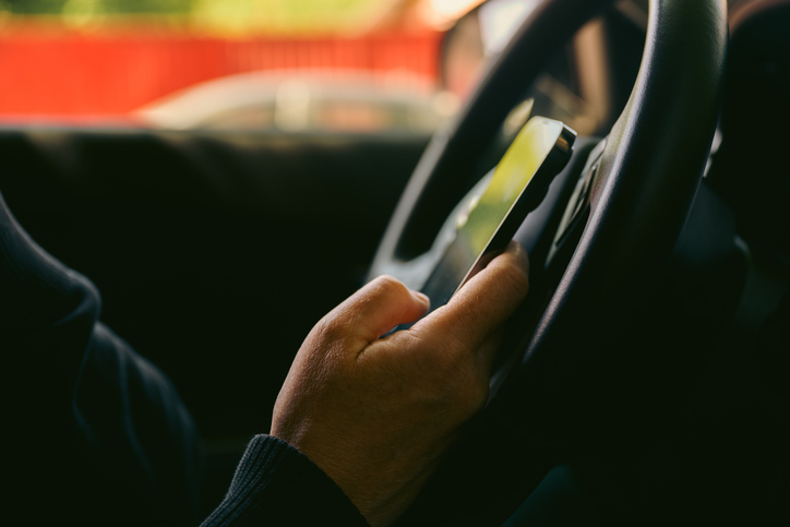 Driver using smartphone while holding steering wheel inside car, distracted driving concept