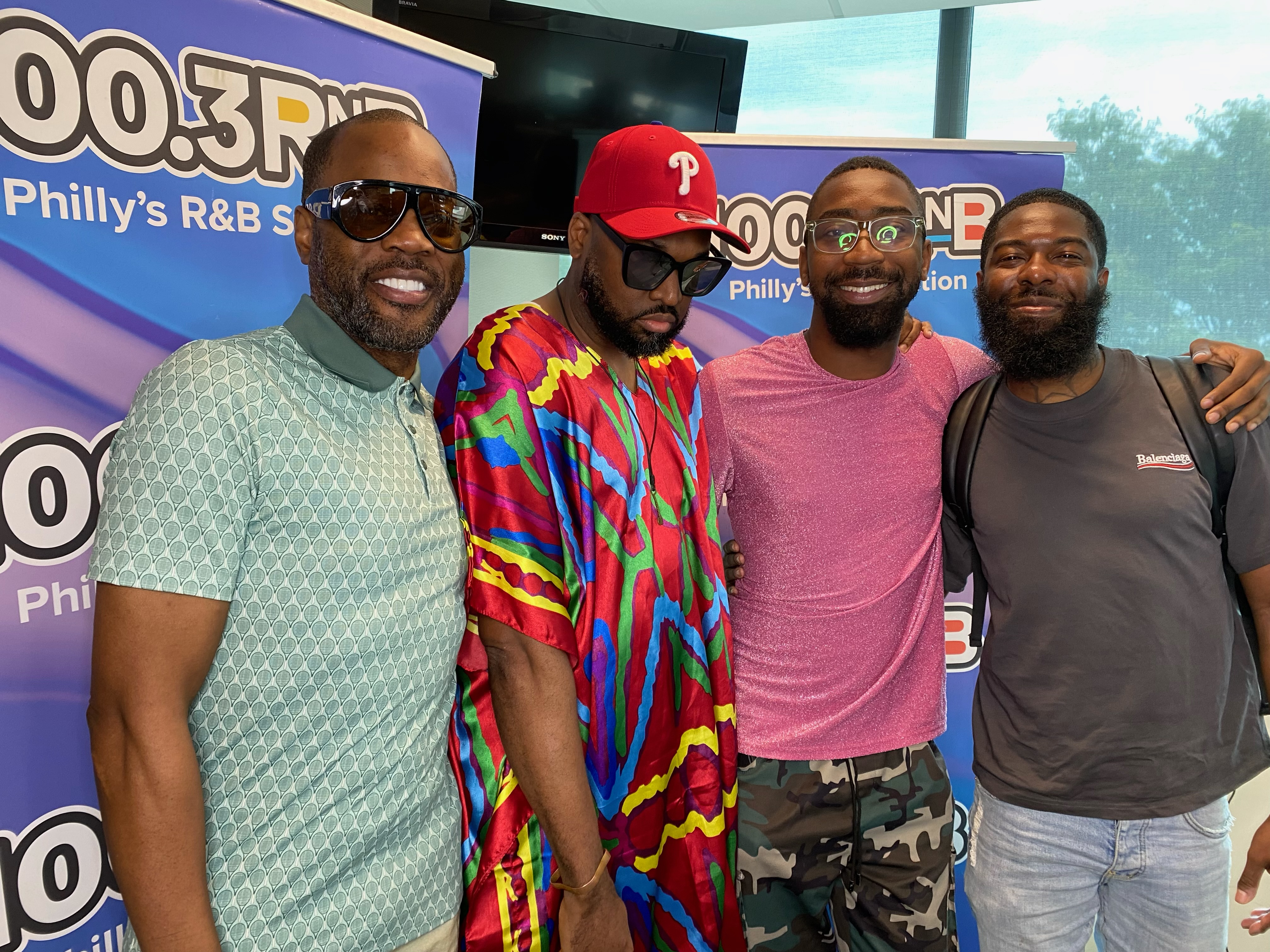 Four men posing together, wearing colorful clothing and sunglasses, in front of a "WDAS 100.3 FM" radio station banner.