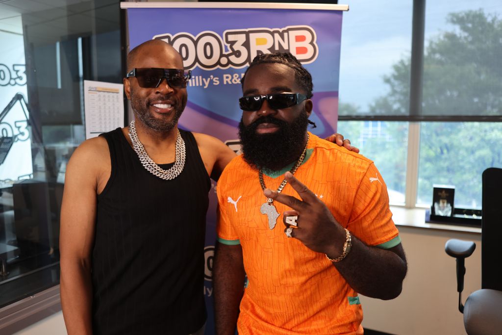 Two men wearing sunglasses and jewelry posing in front of a 100.3 RNB radio station banner.