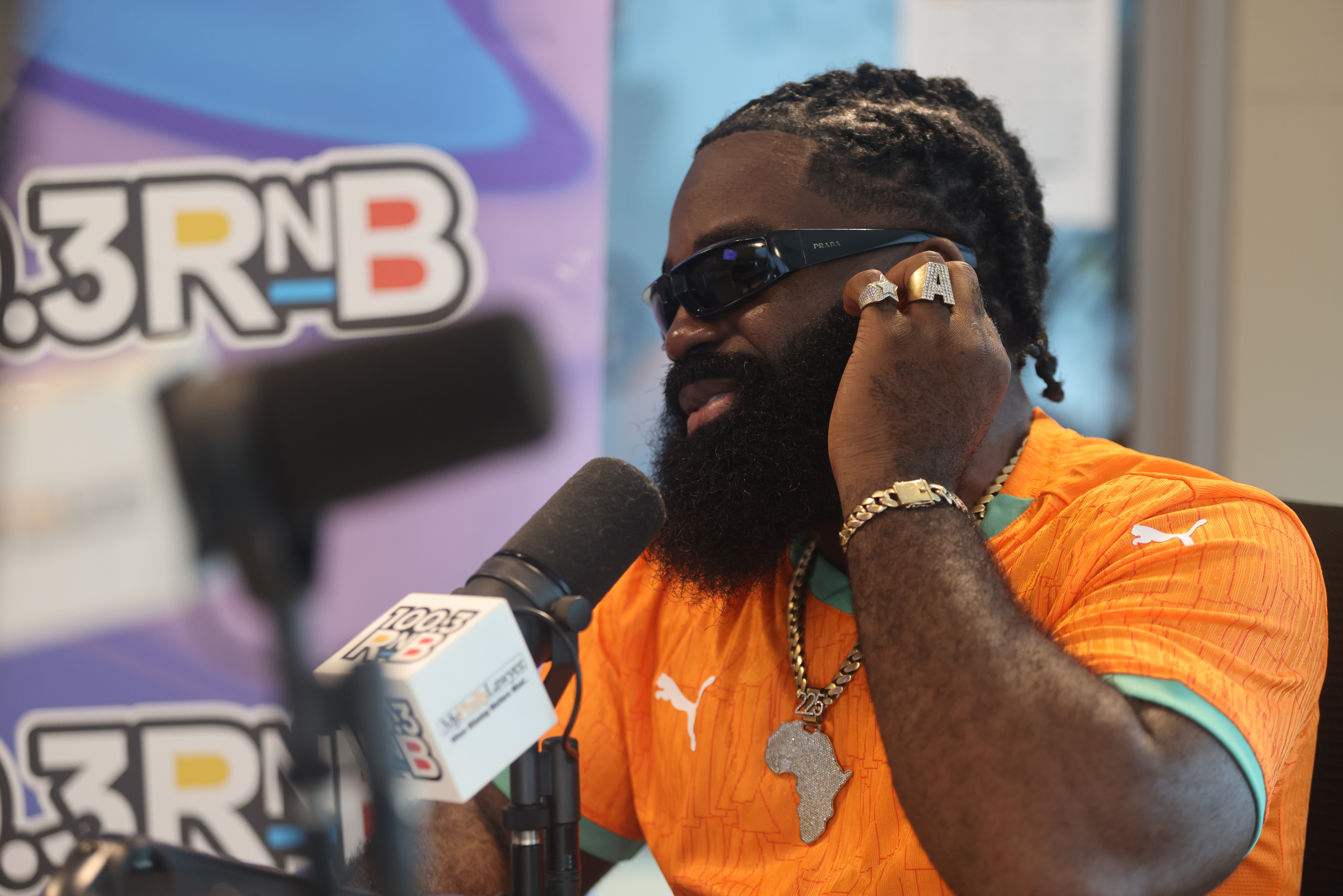 A man with a beard wearing sunglasses and an orange shirt speaking into a microphone in front of a 3RNB logo.