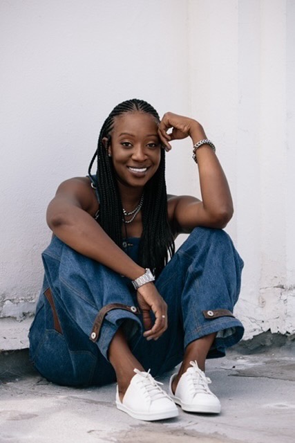 A smiling young woman with braided hair sitting on the ground against a white wall, wearing denim pants and white sneakers.
