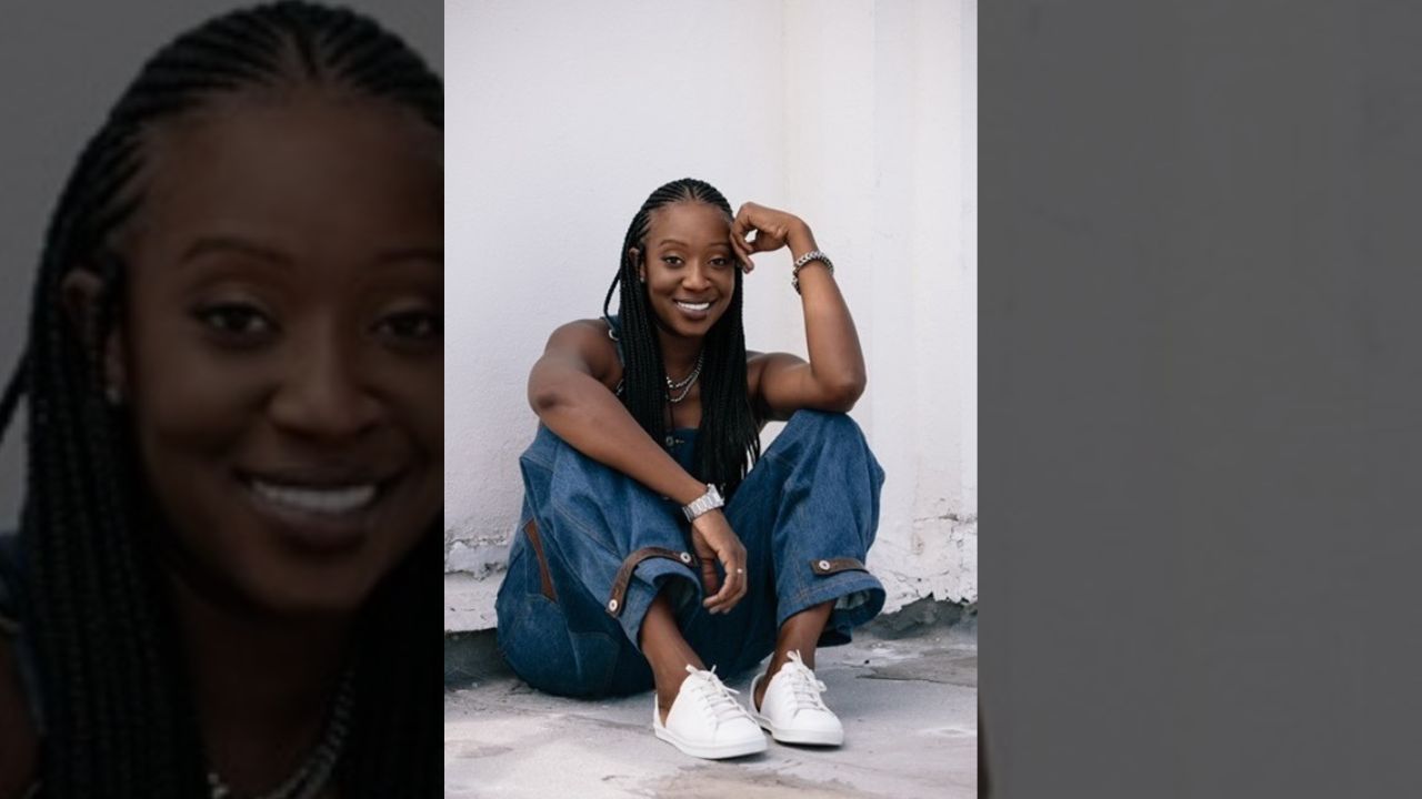 A smiling young woman with braided hair sitting on the ground against a white wall, wearing denim jeans and white sneakers.