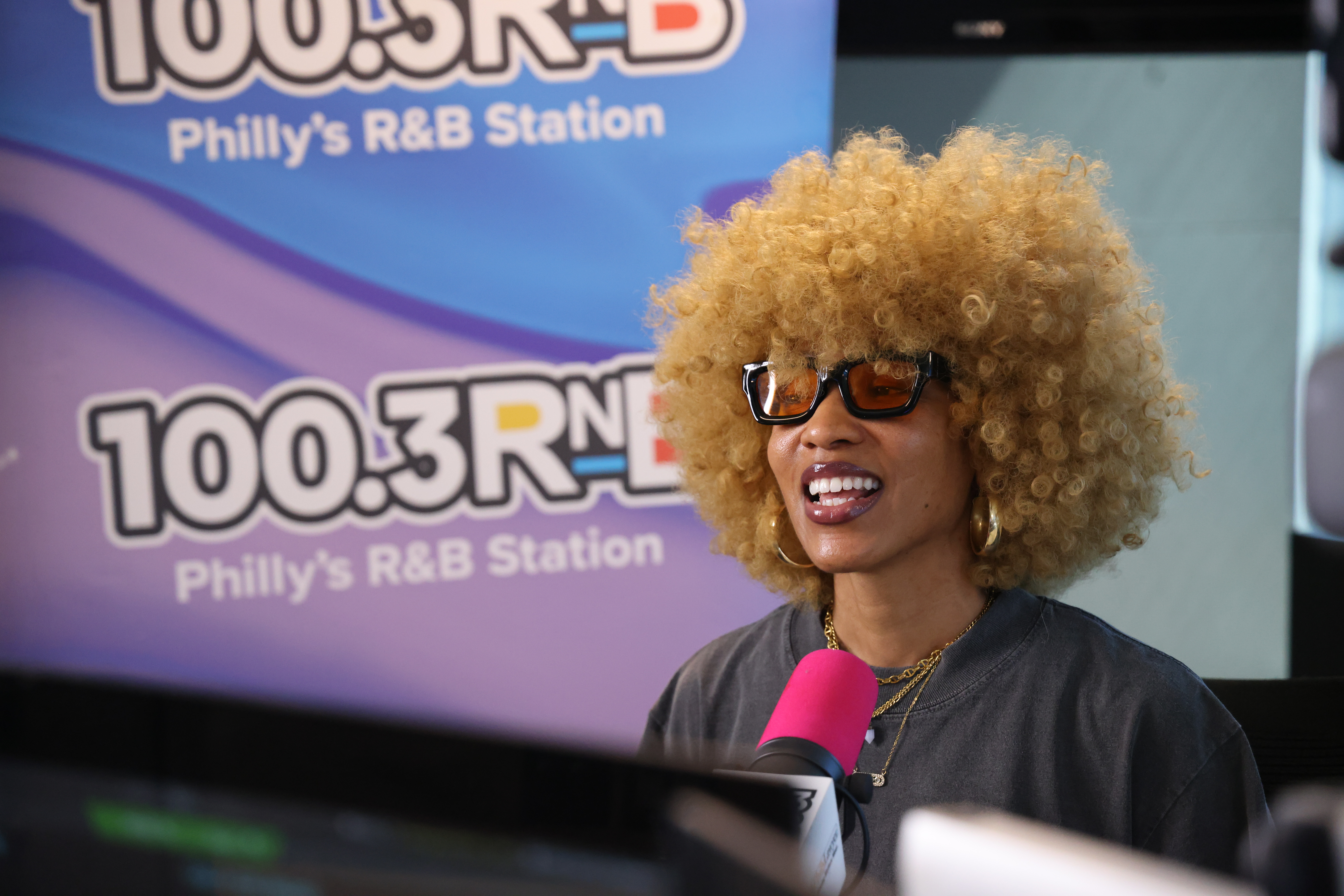 A person with curly blonde hair wearing sunglasses and a pink shirt, standing in front of a banner for "100.3 RNB Philly's R&B Station".