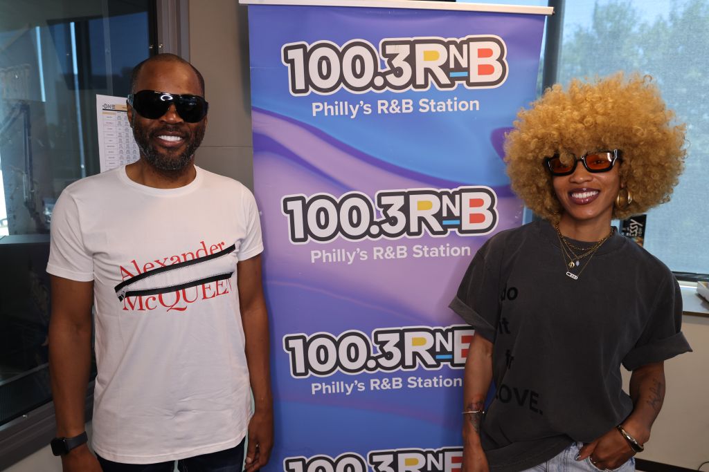 Two people standing in front of a banner for 100.3 RNB, Philly's R&B station. The man is wearing a "Alexander McQueen" shirt and the woman has curly hair and sunglasses.