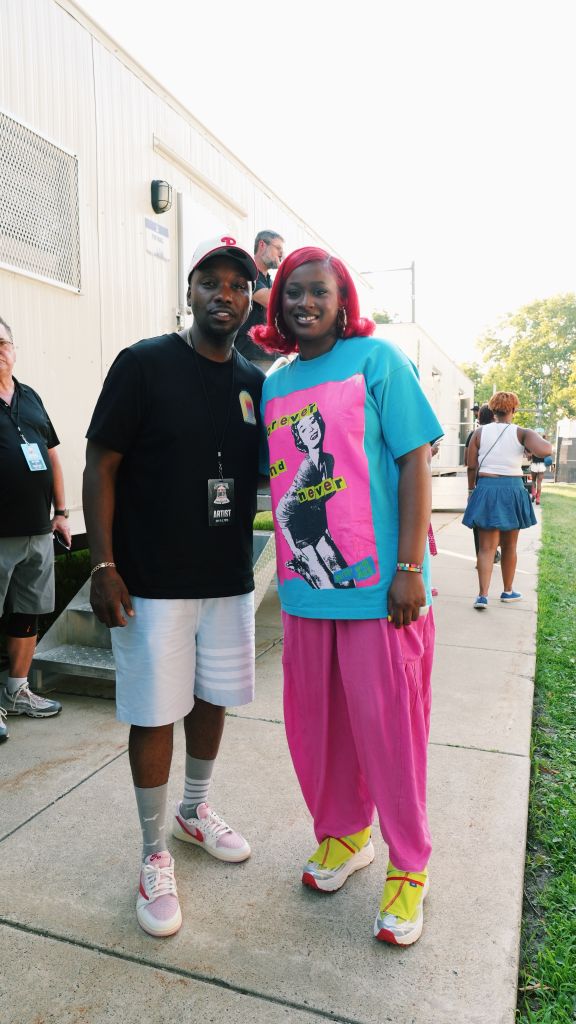 Two people standing together, one wearing a black t-shirt with "ARTIST" text and the other wearing a pink and blue outfit with a graphic t-shirt.