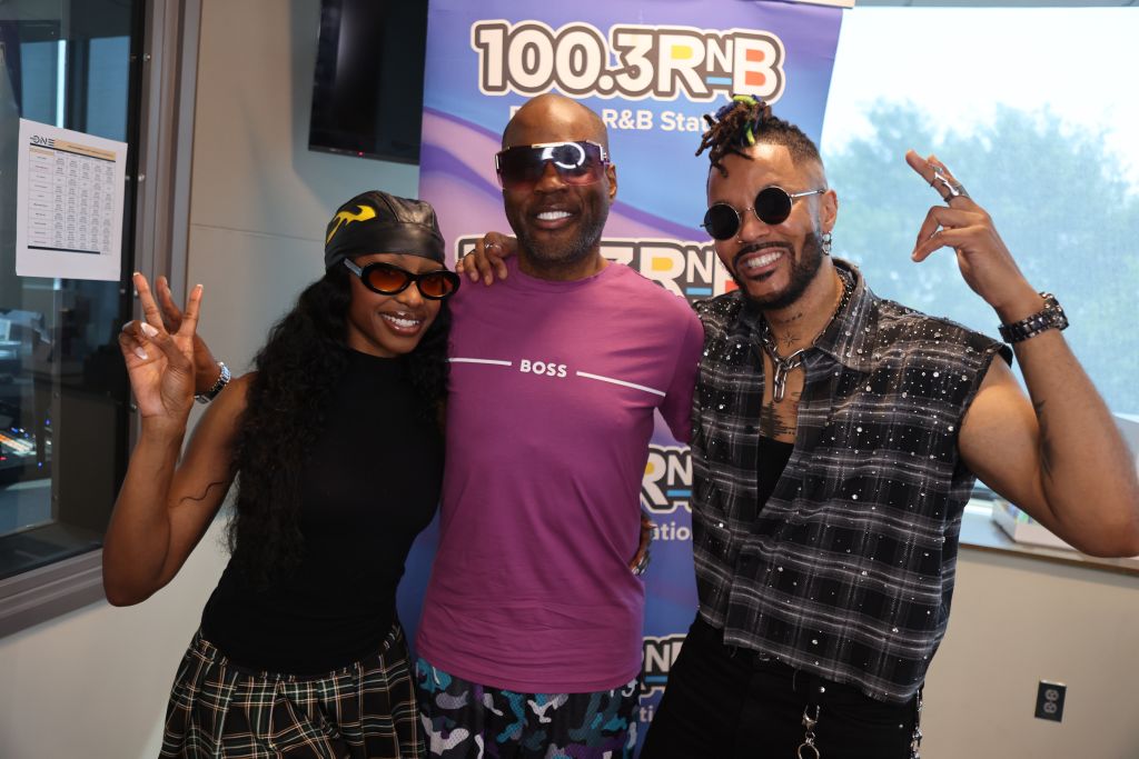 Three people posing together in front of a "100.3 RNB" radio station backdrop, making peace signs and smiling.
