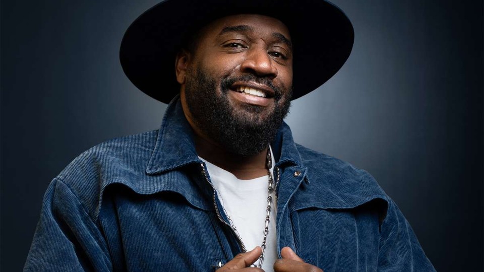 A smiling man with a beard wearing a denim jacket and black hat.
