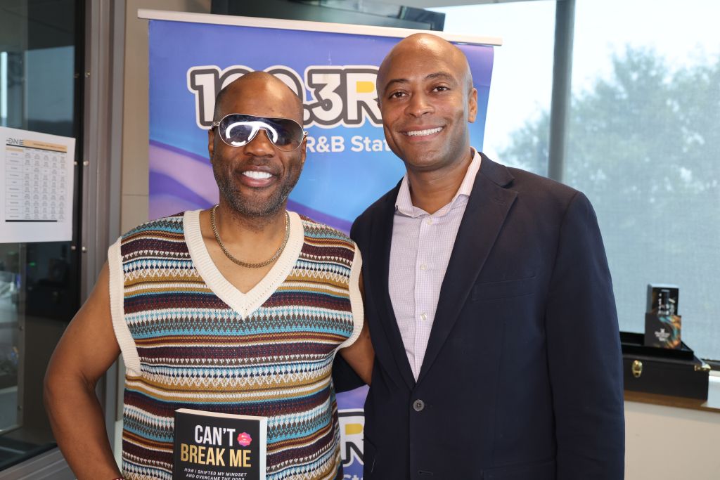 Two men smiling and posing in front of a "103.3 R&B Station" backdrop, one wearing a striped sweater and sunglasses, the other in a suit.