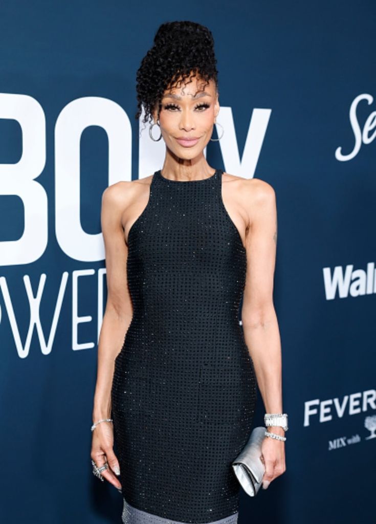 A woman with curly black hair wearing a black sequined dress posing on a red carpet backdrop with "BODY" and "FEVER" text visible.